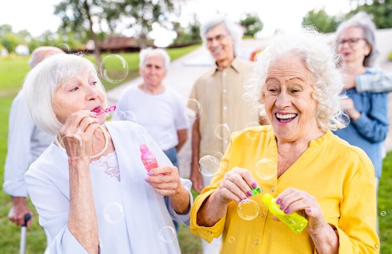 Two Elderly Women Blowing Bubbles With Friends Outdoors at an Independent Living Facility